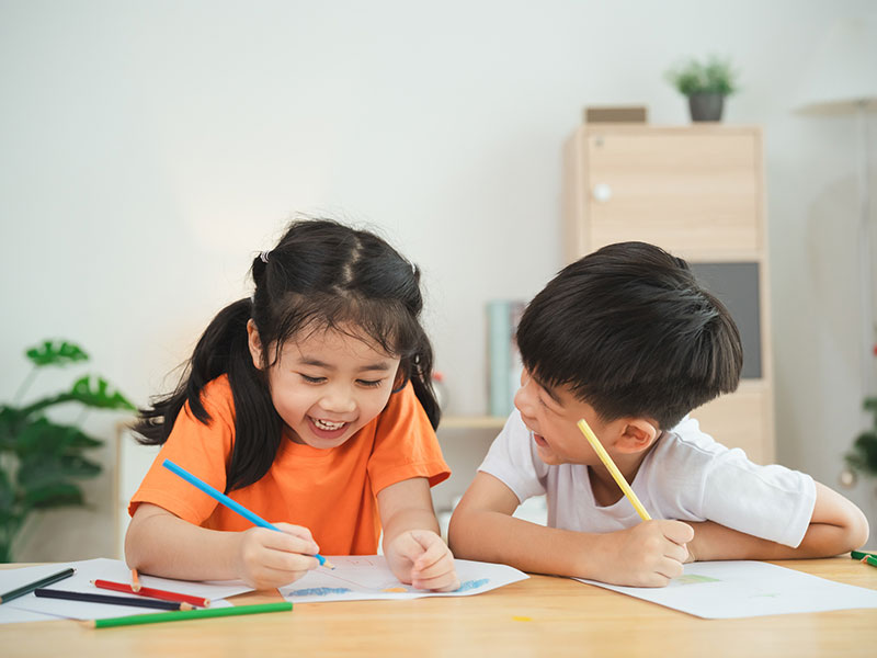 Smiling young girl and boy sitting at a table drawing with colored pencils, enjoying an art activity together indoors.